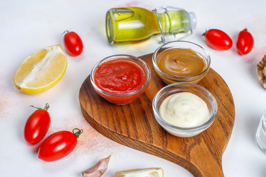 Homemade ketchup, mustard, and mayonaise surrounded by tomatoes, lemon, and olive oil